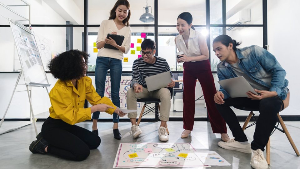 Young creative asian group meeting and looking at project plan lay out on floor discuss or brainstorm business strategy with post note. Workshop for startup team in small office. Happy workplace.