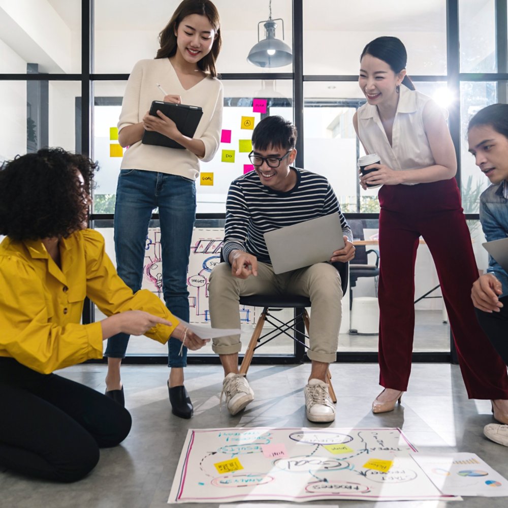 Young creative asian group meeting and looking at project plan lay out on floor discuss or brainstorm business strategy with post note. Workshop for startup team in small office. Happy workplace.