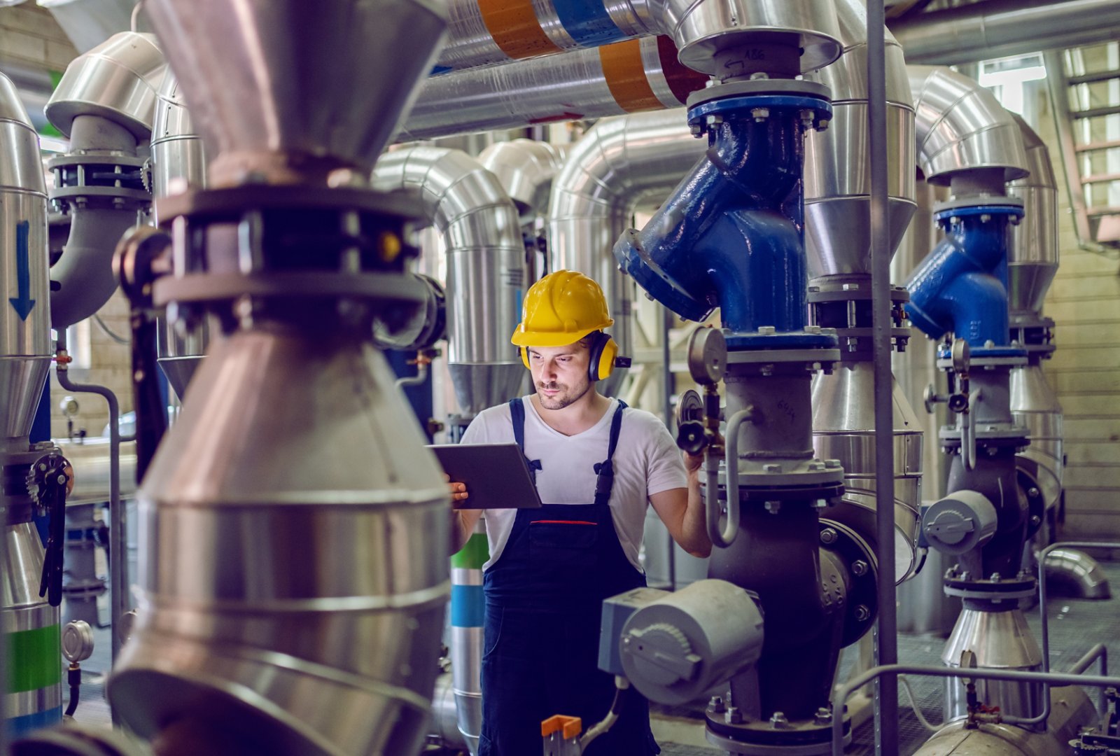 Handsome serious caucasian worker in overalls, with protective helmet and antiphons adjusting pressure on boiler while holding tablet and standing in factory.