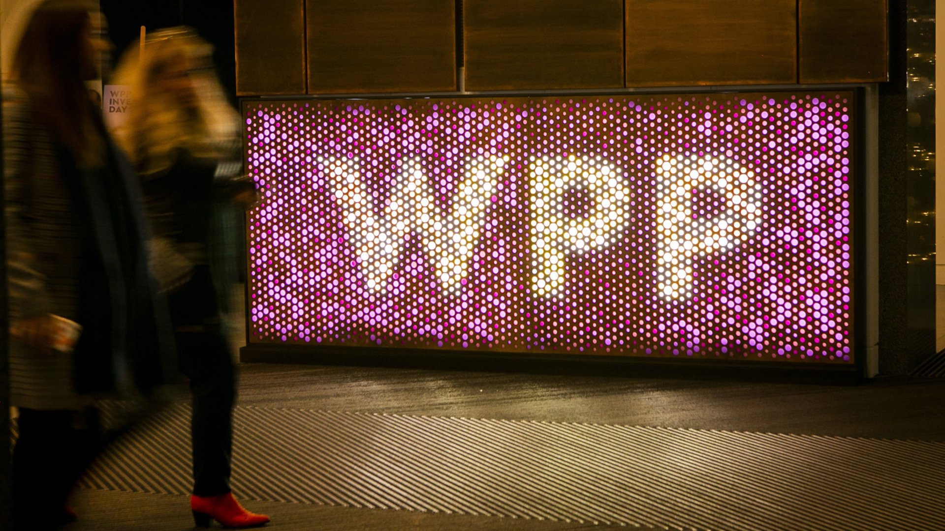 A large illuminated wall displays the letters WPP formed by numerous circular lights. The sign glows in shades of pink, purple, and white, creating a vibrant focal point in the scene. Blurred pedestrians walk past in the foreground, suggesting motion and an urban environment. The overall atmosphere is modern and dynamic, with strong nighttime lighting and reflections on the ground.