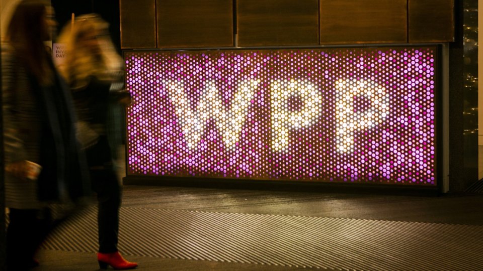 A large illuminated wall displays the letters WPP formed by numerous circular lights. The sign glows in shades of pink, purple, and white, creating a vibrant focal point in the scene. Blurred pedestrians walk past in the foreground, suggesting motion and an urban environment. The overall atmosphere is modern and dynamic, with strong nighttime lighting and reflections on the ground.