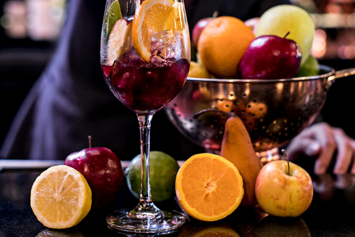 A man pours red liquid into a wineglass full of orange, apple and lime slices while fresh fruits rest on the counter