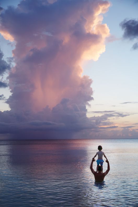 A man holding a young boy on his shoulders in waist deep ocean water looking at purple clouds.