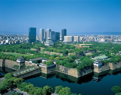 Fortress-like structure with dense foliage, all hemmed in by the river in the foreground and the city in the background