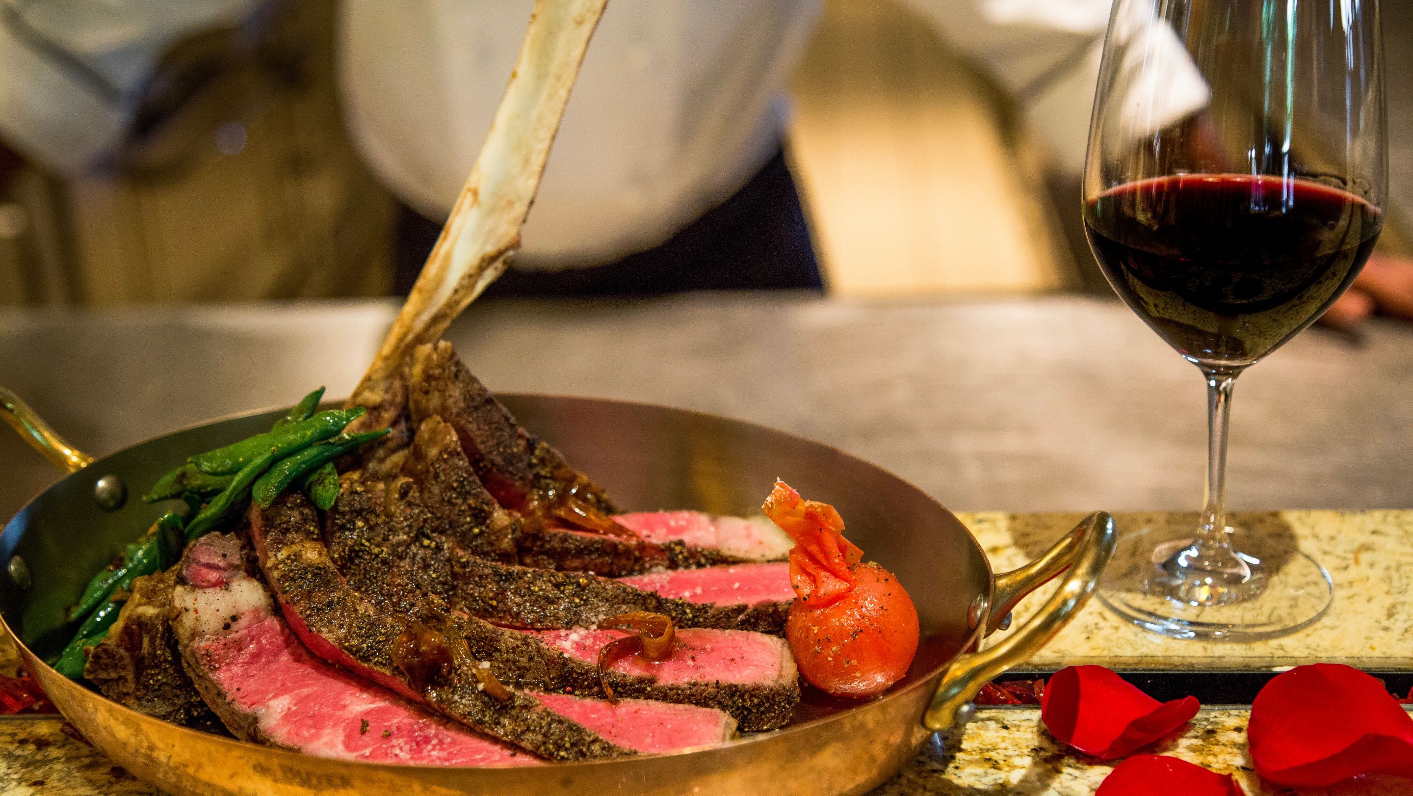 A chef stands over a presentation of a steak and glass of wine
