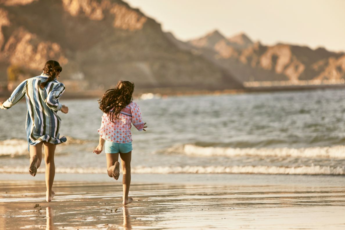 Two children running along the wet sands of a beach with rocky mountains in the background.