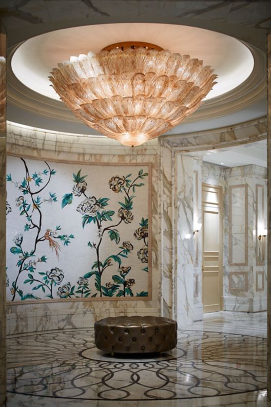 A marble hallway with a round tufted bench underneath a large chandelier and in front of a floral mural.