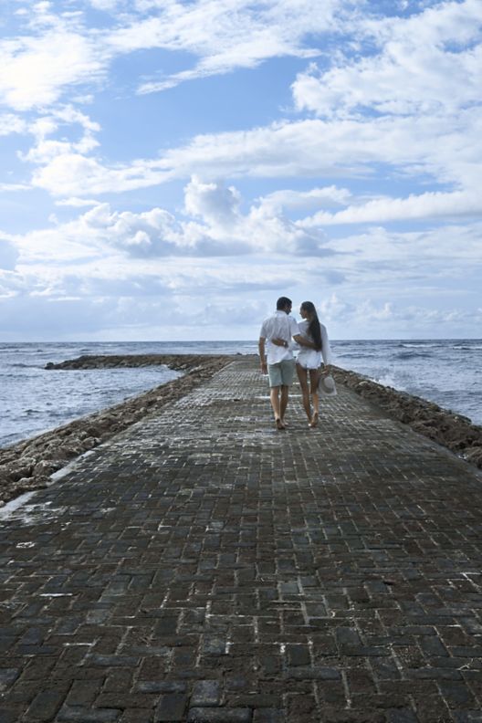 A couple with their arms around each other walking down a paved walkway surrounded by the ocean