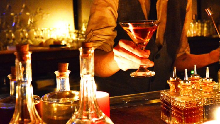 Bartender holds a cocktail in a martini glass over a bar set with multiple glass bottles