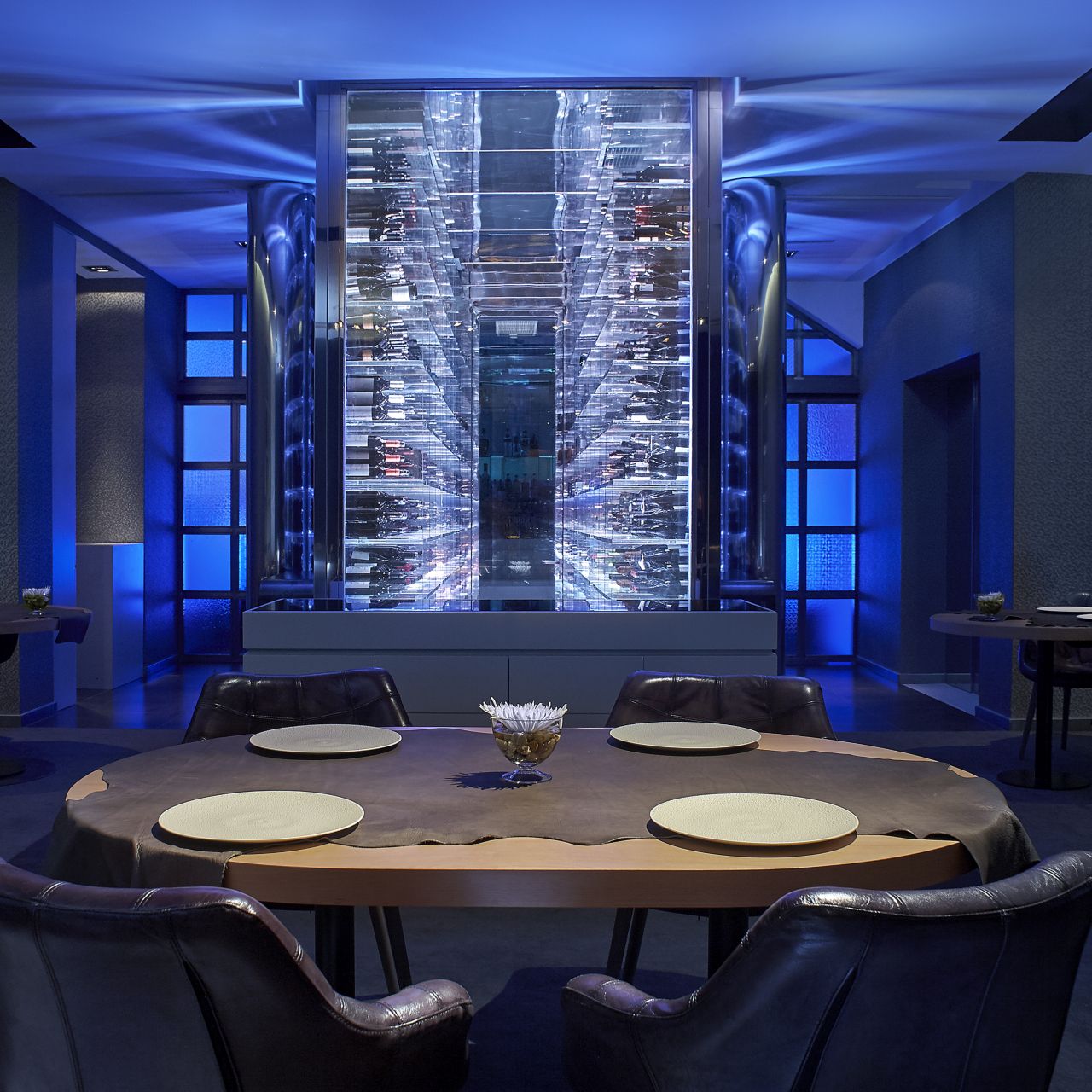 A dining table in front of a large glass wine closet backlit for the evening