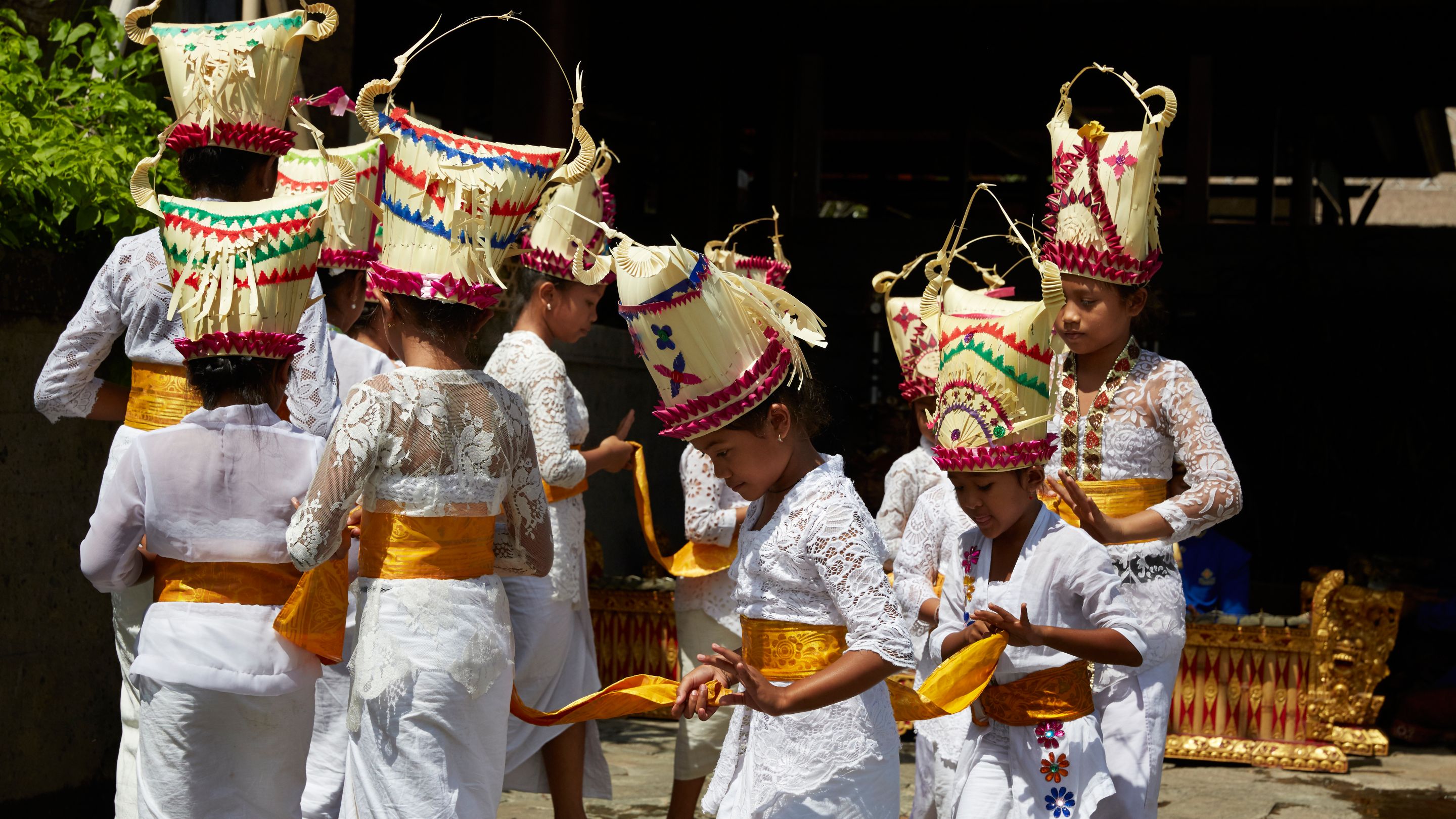 Children wearing white gowns with golden sashes and decorative headdresses walk in line