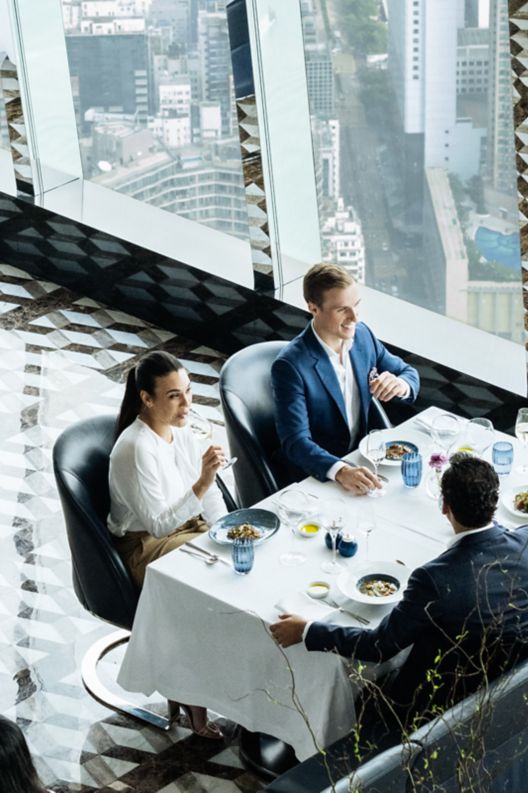 People sitting at tables with white table cloths and view of city skyline out of large windows.