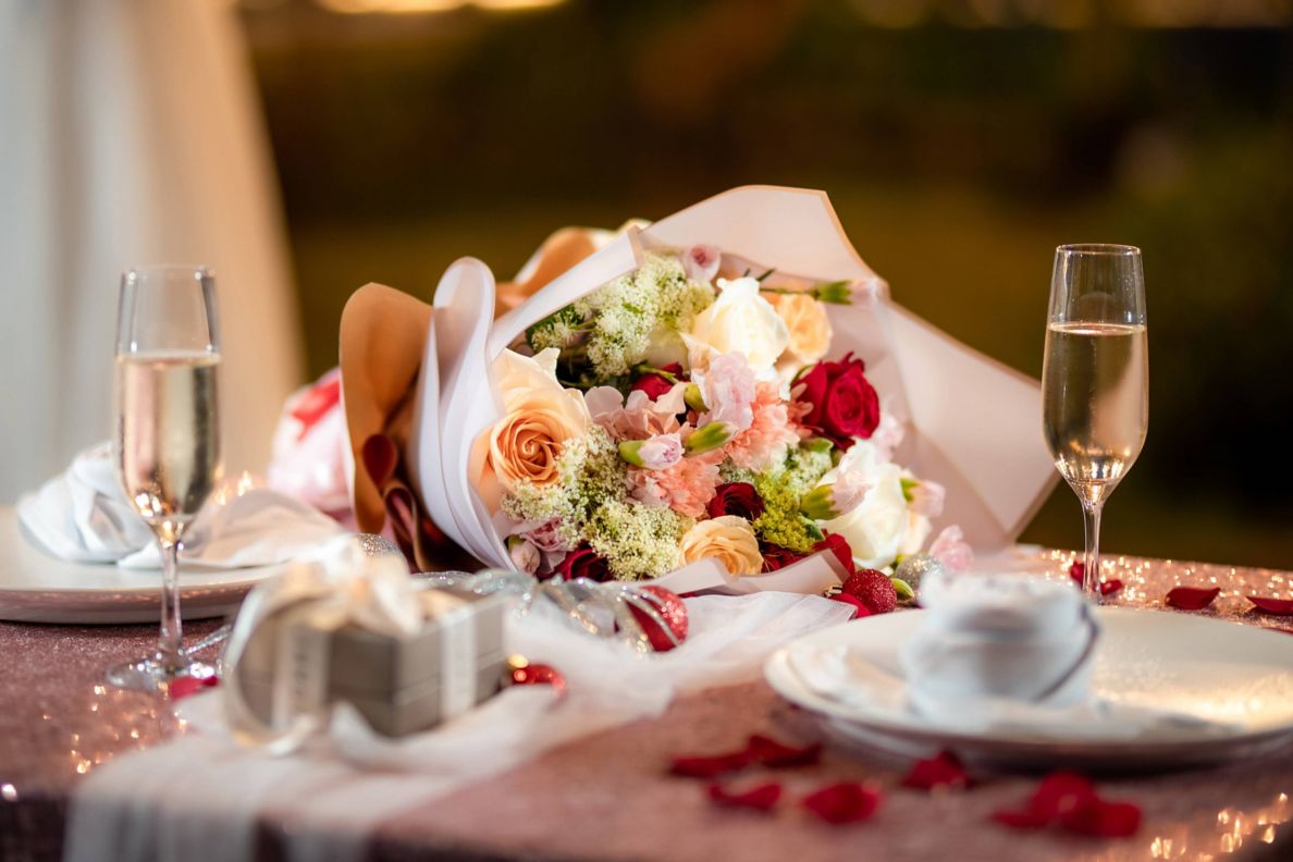 A dining table with a bouquet of flowers, champagne flutes, an engagement ring box and rose petals on top.