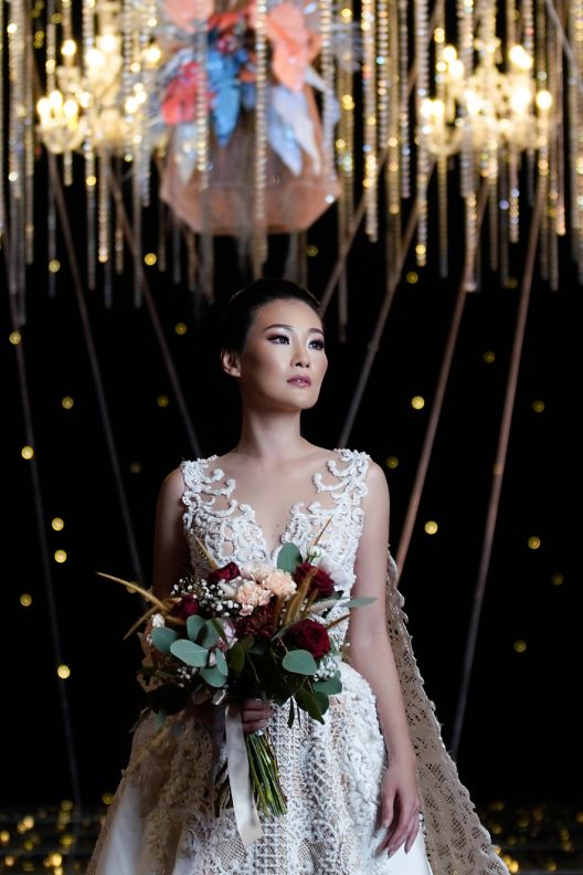 Bride standing under hanging chandelier lights while holding a bouquet of flowers.