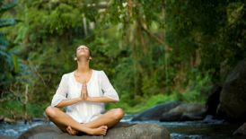 Woman in a white tunic sits cross-legged on a boulder whilst meditating in the rainforest
