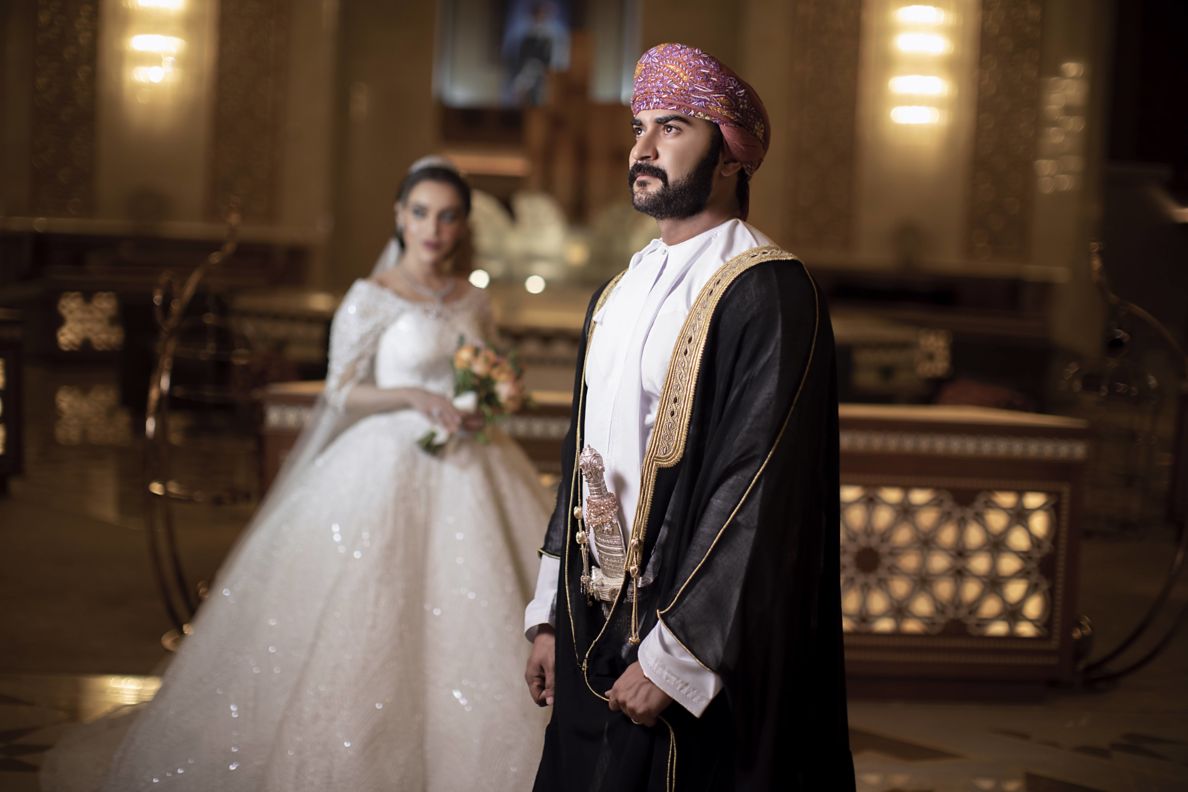A groom in traditional, formal, Omani garments stands inside the Atrium Lounge with is white gown clad bride in the background.
