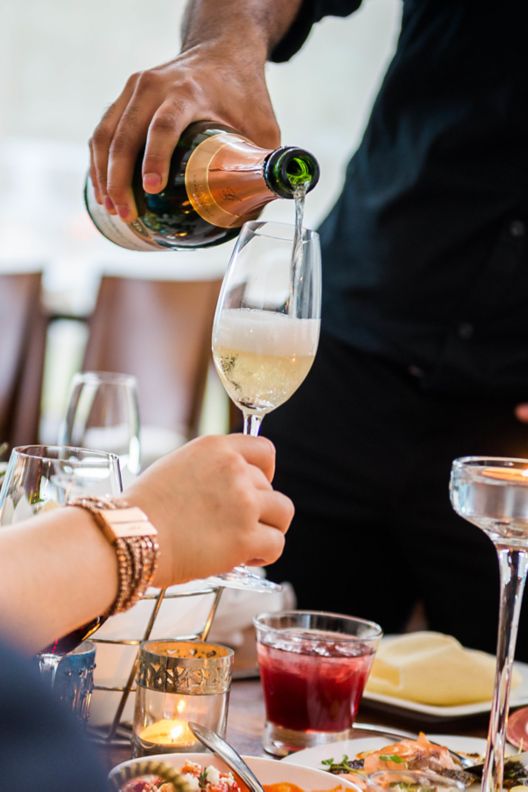 Someone pouring champagne into a glass above a dining table with plates of food on top.