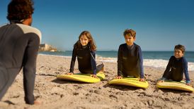 Three kids lie on surfboards on the sand while an instructor shows them correct form