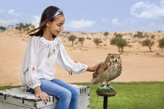 A young girl in jeans and a white peasant top sits outside while smilingly petting a large brown-and-white speckled bird
