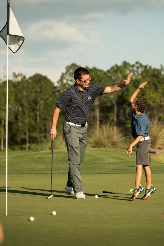 A young boy giving his golf instructor a high five with two young golfer friends in the foreground.