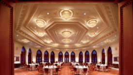 Geometric ballroom with arched windows lining the walls, ornate ceiling detail, chandeliers and tables on red carpet