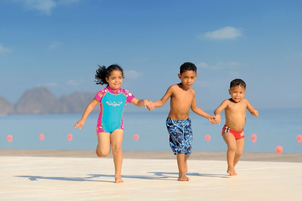 Three kinds holding hands on the beach with the sea and mountains in the background.