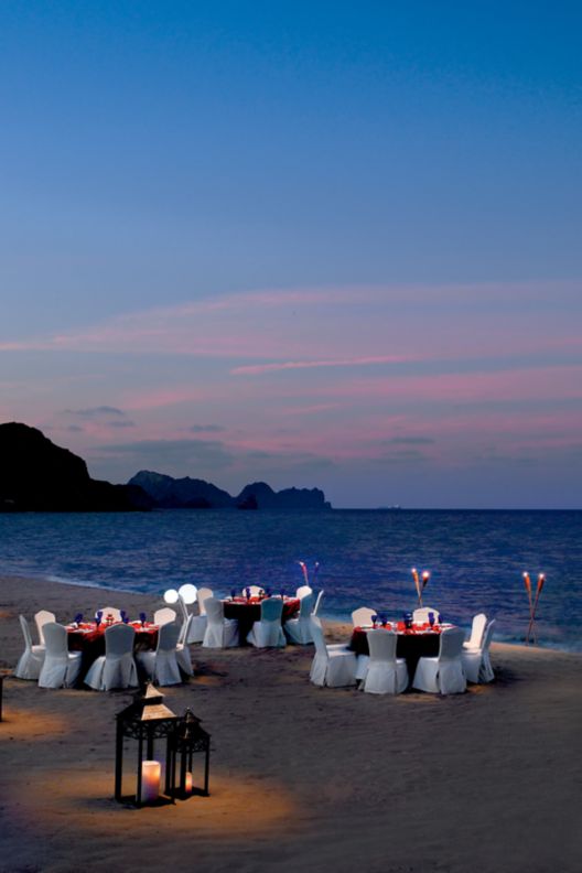 Dining tables and chairs illuminated by candles arranged on a beach near the sea at dusk with mountains in the background.