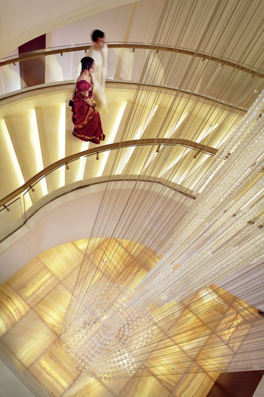 Couple in traditional Indian wedding attire walk along a curvaceous staircase suspended next to a crystal light fixture.
