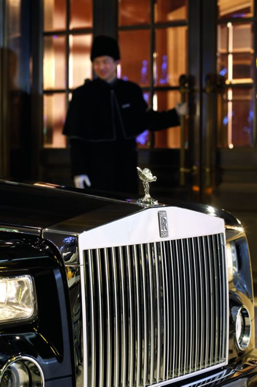 A luxury car pulls up to the front of a building where a doorman holds the door