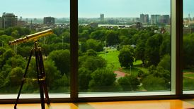 A telescope overlooks a lush green park with tall buildings in the distance