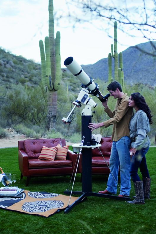 Two people using a telescope to stargaze with a couple arm chairs behind them.