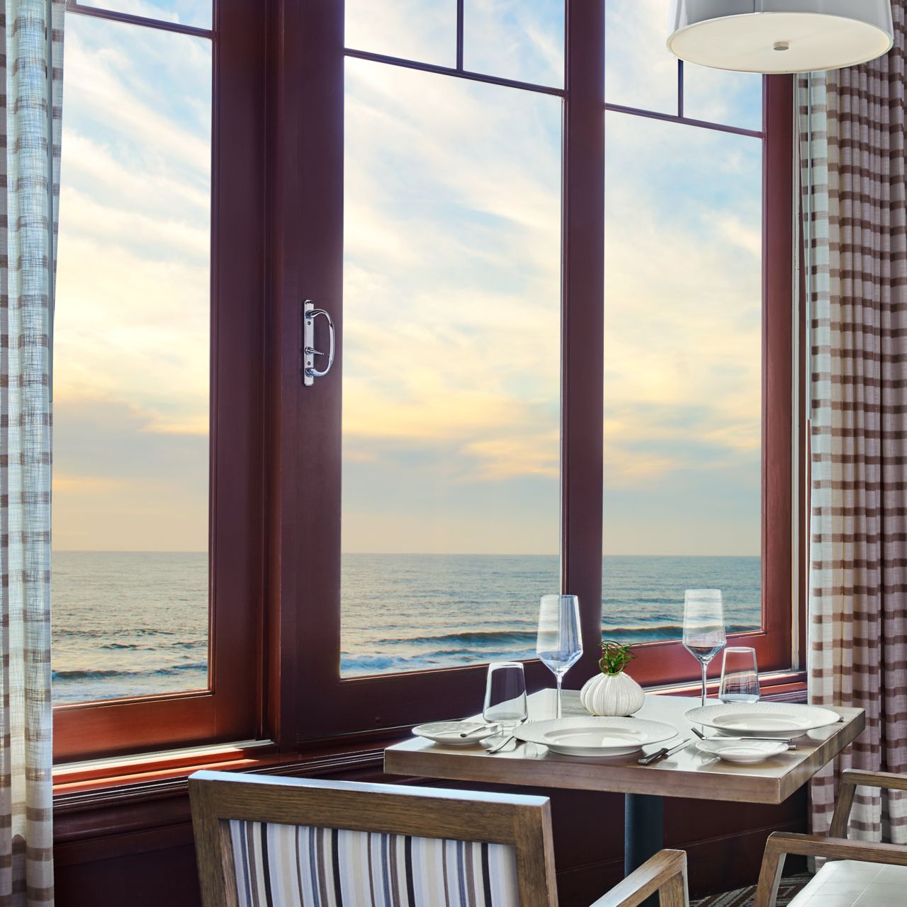 Square table with two armchairs, wine and a plate of food next to a window overlooking the ocean