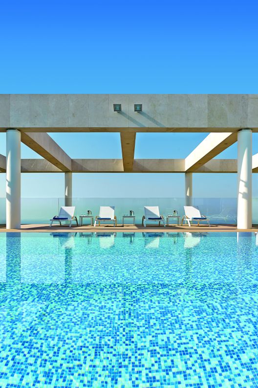 Aquamarine rooftop pool leads to four lounge chairs with a glass railing behind them and views of the Mediterranean beyond