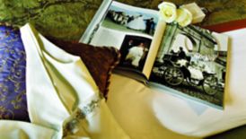 A white sleeveless gown, two white roses and an open photography book with wedding imagery all scattered on a bed