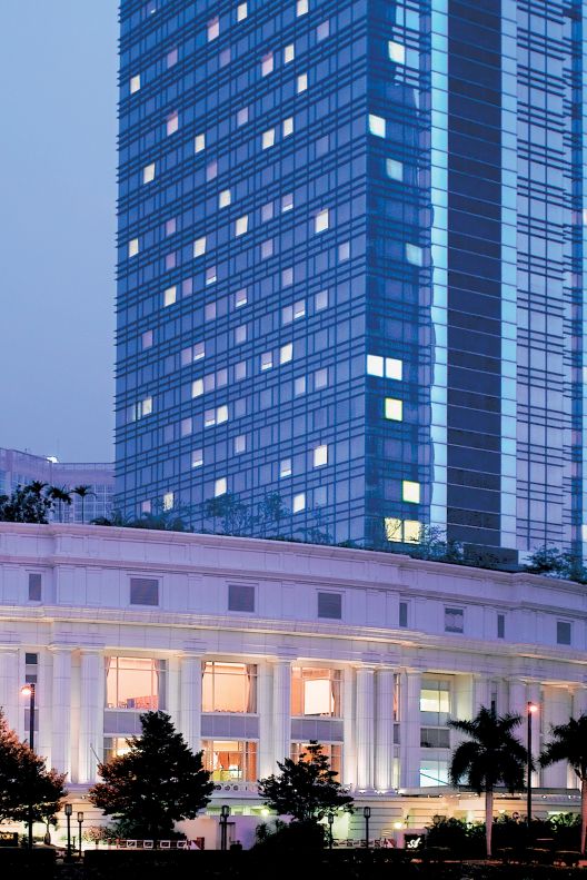 A modern skyscraper soars upwards behind the hotel?s elegant and traditional white façade as dusk settles over the city