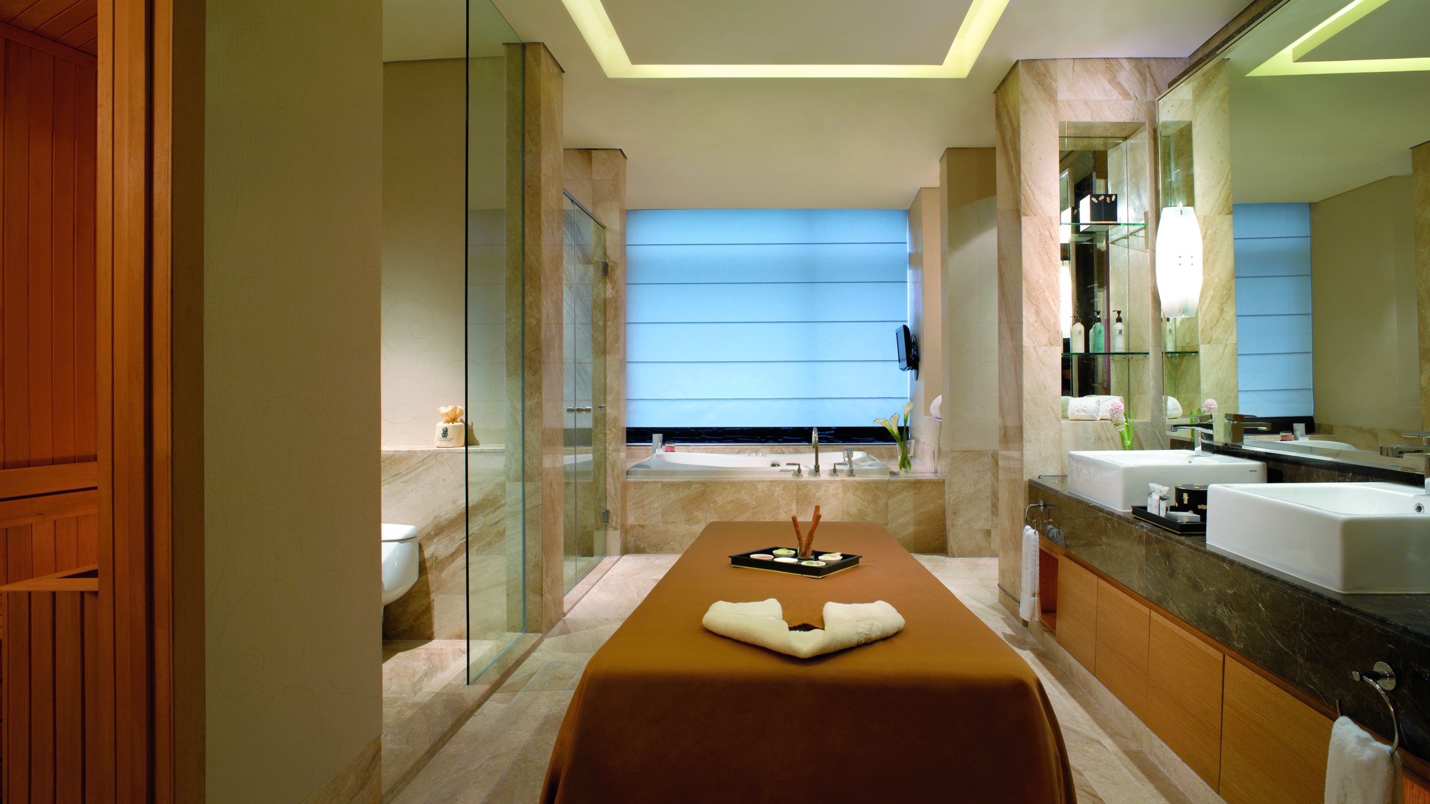 Massage table in the middle of a marble bathroom with a double vanity, glass-enclosed shower and large tub
