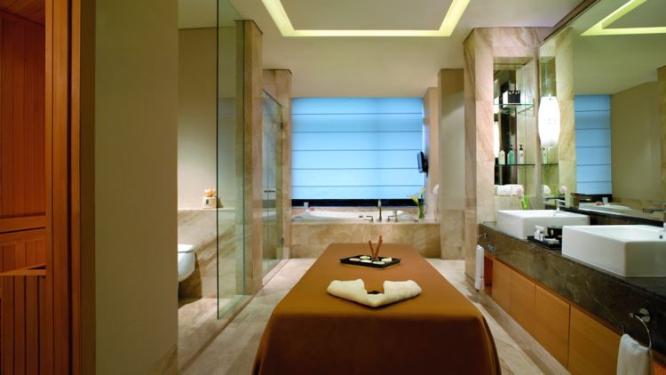 Massage table in the middle of a marble bathroom with a double vanity, glass-enclosed shower and large tub