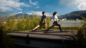 A man and woman in black workout clothes stretch on an open-air platform overlooking the mountains