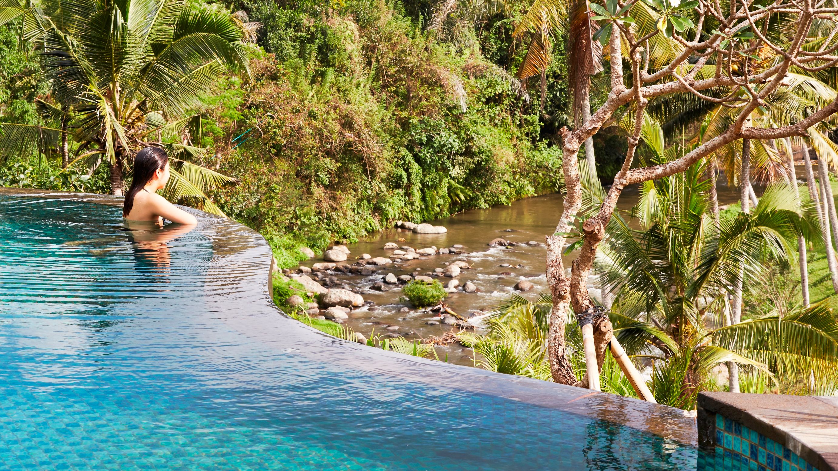 一位女士坐在蓝绿色礁湖风格泳池边眺望河流，俯瞰雨林景观