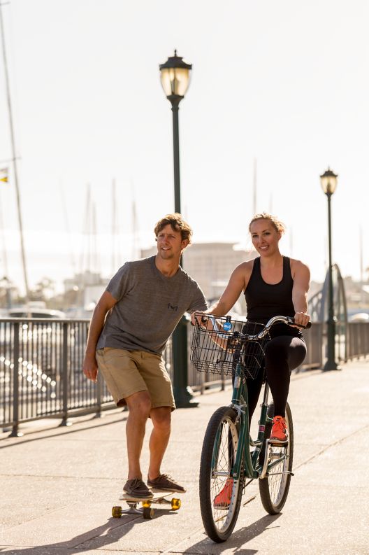 A skateboarder and a bicyclist travel down a marina boardwalk
