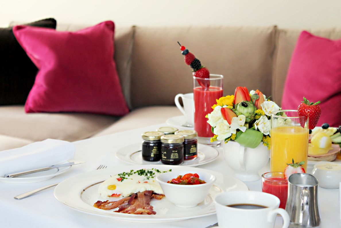 Breakfast spread with bacon, eggs, fruit and juice with a couch in the background.