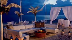 Hurricane lamps illuminate white couches, palm trees and drapes during an evening reception on the beach