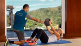 A woman holding a medicine ball does abdominal crunches with the help of a male trainer