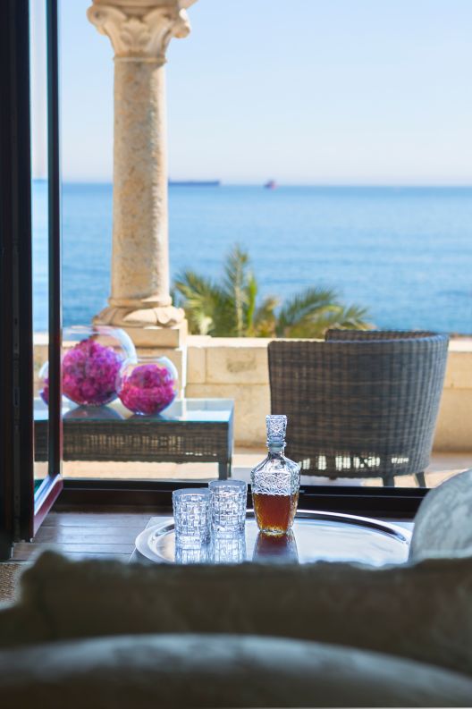 A seating area with a carafe of alcohol and glasses on a table overlooks a furnished patio facing the sea