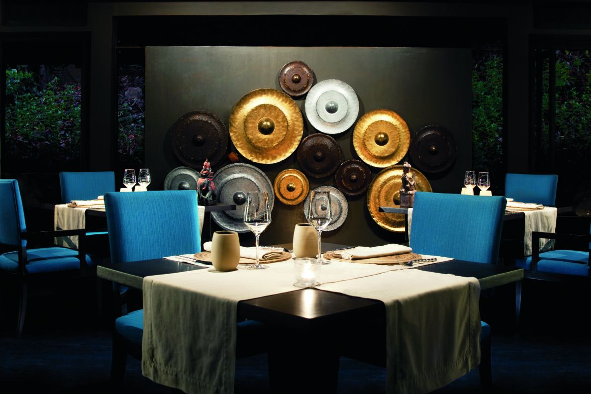 Three square dining tables underneath a chandelier and next to a wall with a plate-like art installation