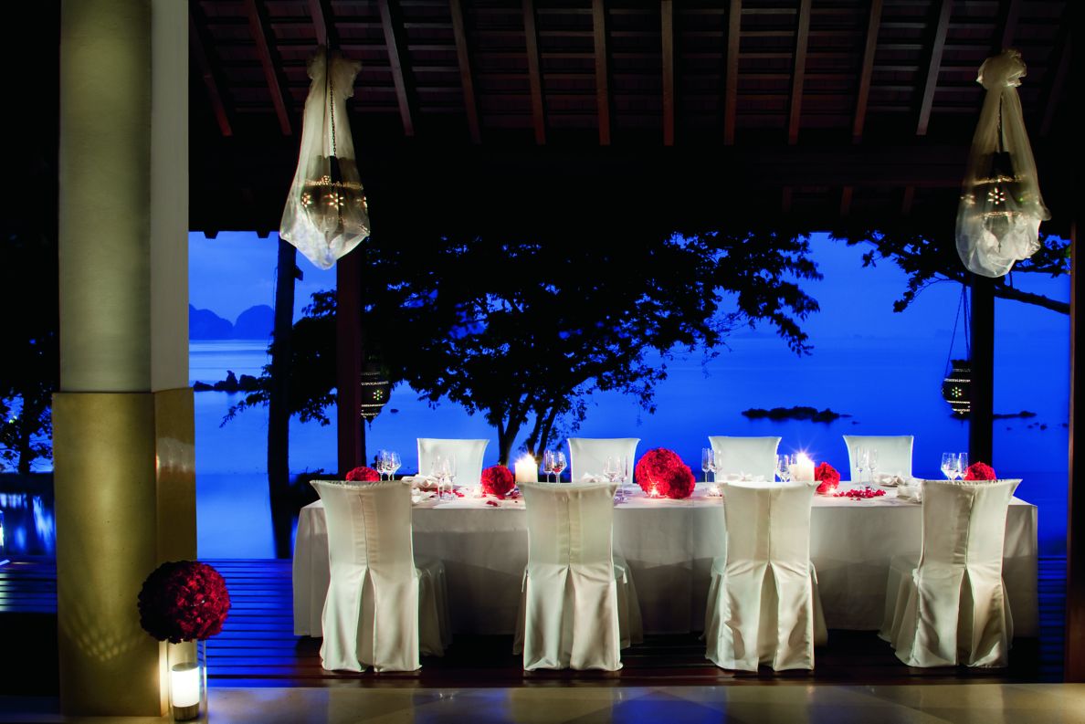 夕日を背景に、テラスに、白いスリップカバー付きの椅子、白いテーブルクロス、赤い花のセンターピースを配した8人用のテーブルセッティング