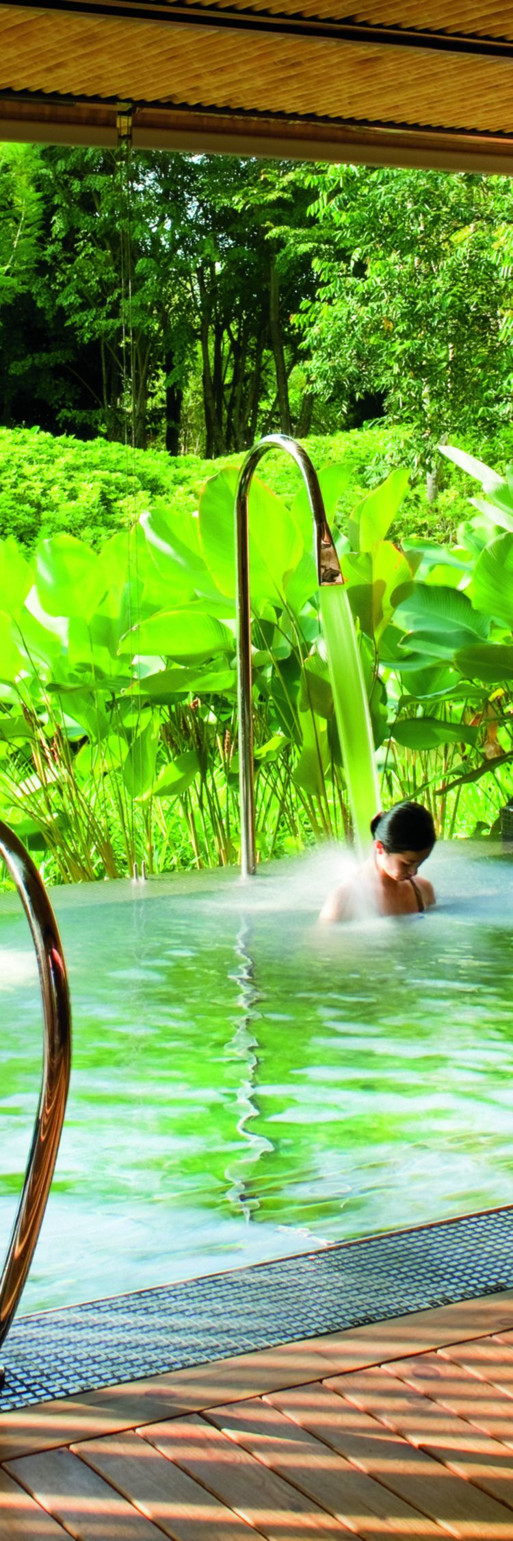 Woman sitting under a jet of water within a square vitality pool located in a semi-enclosed room that opens onto the garden