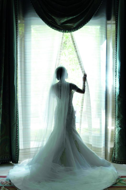 A bride gazes out a floor-to-ceiling window while dressed in a mermaid-style gown and veil