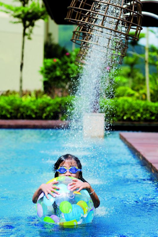 A young girl holding on to an inflated ball in an outdoor pool under a shower of water.