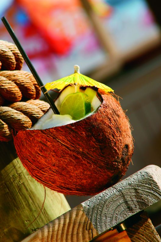 Coconut on wood stool with a straw, sliced cucumber and drink umbrella inside of it. 